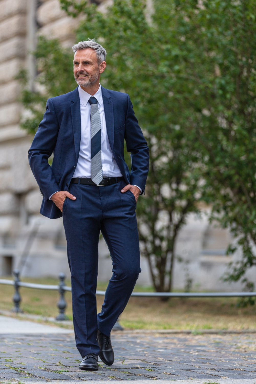 Egy ősz hajú és szakállú férfi magabiztosan lép ki, elegáns ZAQQ BRIQ Captoe Black cipőt viselve. Sötétkék öltönnyel, fehér inggel és csíkos nyakkendővel kombinálja őket. Egyik kezét zsebre téve sétál el egy épület és fák mellett.