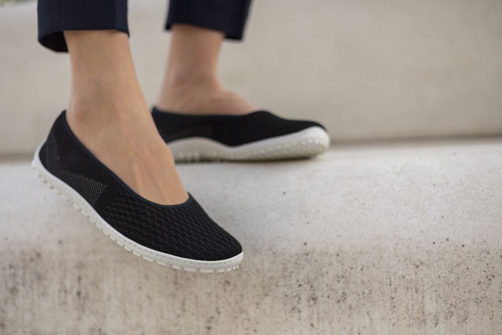 Egy személy SUQAR Black papucsot visel a ZAQQ-tól, sima hálós textúrával és fehér sneaker talppal, és betonlépcsőkön áll. A fókusz művészien megörökíti a lábfejét ezekben a papucsokban, valamint a sötét nadrágba bújtatott lábszárának egy részét, így ötvözve a kényelmet a finom nőies megjelenéssel.