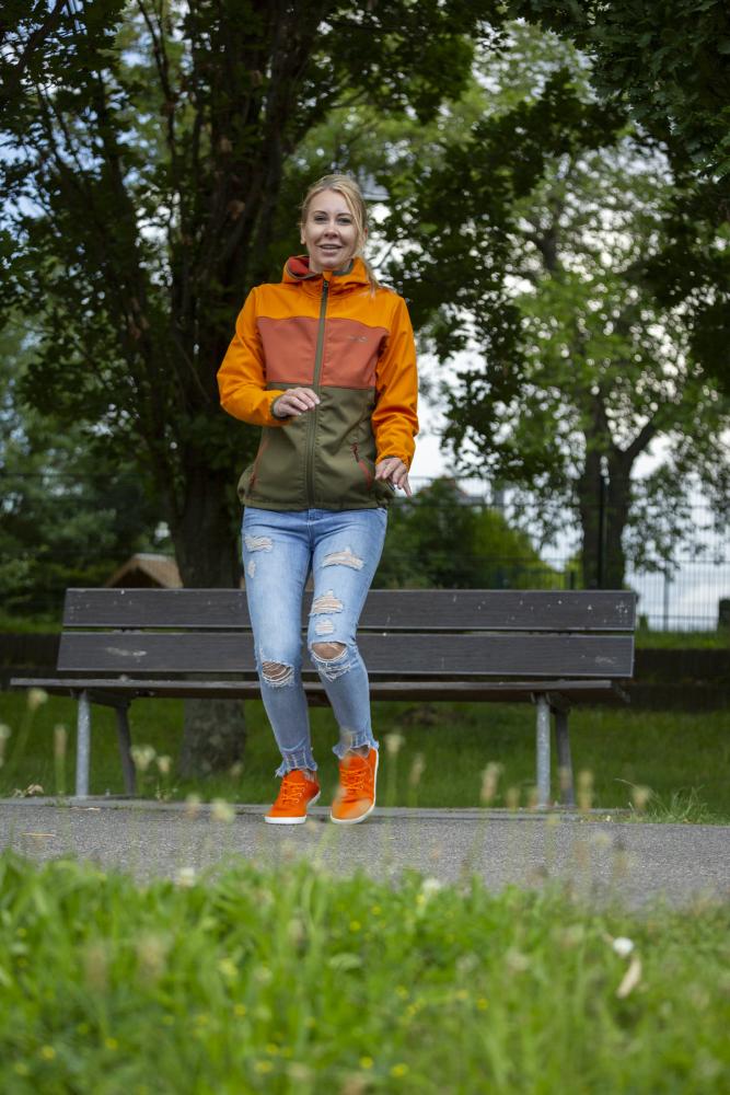 Egy nő narancs-zöld kabátban és szakadt farmerben sétál egy parkösvényen, és élvezi a mezítlábas érzést, amelyet élénk narancssárga ZAQQ LIQE cipői nyújtanak. Mosolyogva halad el egy fapad és zöld fák mellett a felhős ég alatt.