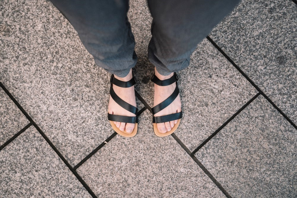 Egy személy lábaira nézve a ZAQQ NIQ Black szandálokban, kiváló minőségű nappa bőrből, akik egy kőlapokkal borított járdán állnak. Az elegáns fekete pántos dizájn maximális kényelmet nyújt és tökéletesen kiegészíti a sötét nadrágot.