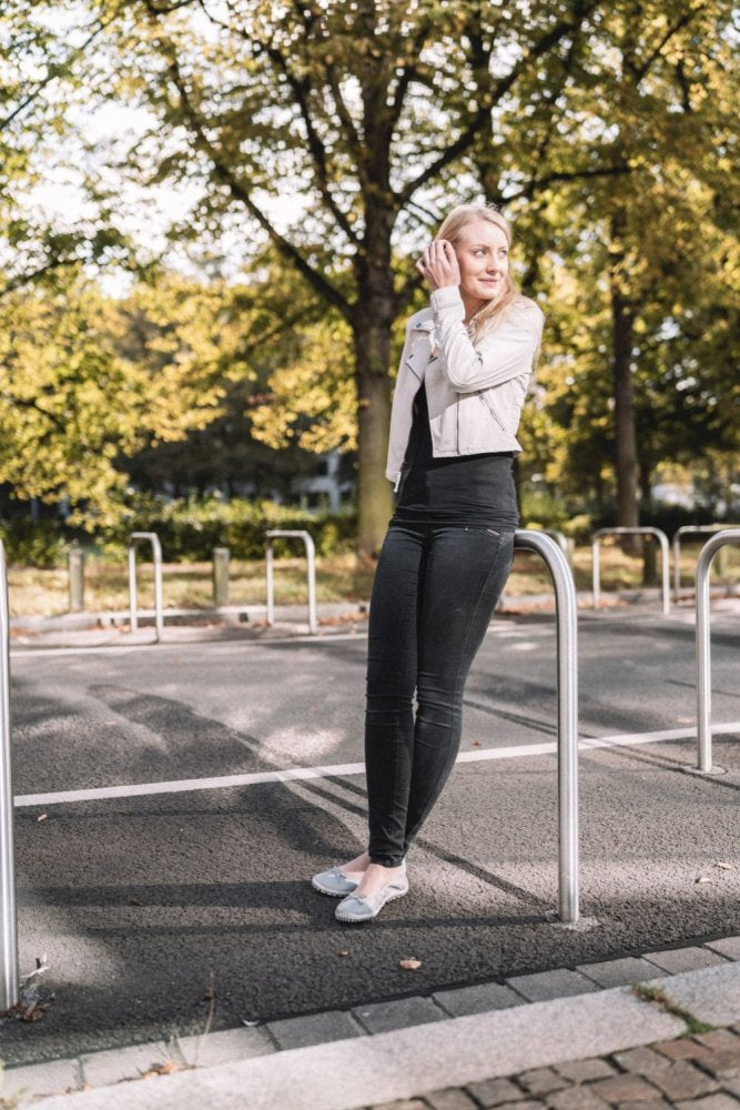 Egy hosszú szőke hajú személy, aki fehér kabátot és sötét farmert visel, ZAQQ SUQAR Grey cipőkkel kiegészítve, egy utcán áll és egy fémoszlopnak támaszkodik. A háttérben zöld levelű fák láthatók, és a napfény átszűrődik az ágakon.