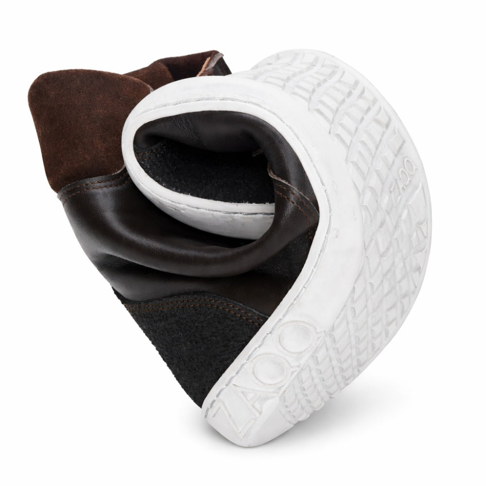 Der MINSQ Dark Brown Sneaker von ZAQQ aus schwarzem Leder mit mittelhohem Schnitt lässt sich in der Mitte biegen, um seine Flexibilität zu demonstrieren. Seine weiße Gummisohle weist ein strukturiertes Muster auf und zeigt deutlich den Markennamen ZAQQ. Dieser Barfußschuh-Stil verfügt außerdem über eine Aufreißlasche auf der Rückseite.