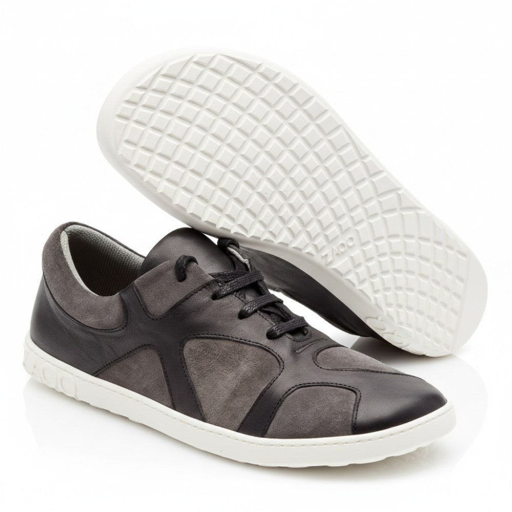 Ein Paar schwarze QAANAAQ-Freizeitsneaker mit flexiblem Design und strukturierter Sohle aus leichtem Veloursleder. Der Sneaker rechts steht aufrecht, während der linke geneigt ist, um die Sohle hervorzuheben. Sie werden mit schwarzen Schnürsenkeln geliefert und tragen den Markennamen ZAQQ deutlich sichtbar an der Seite.